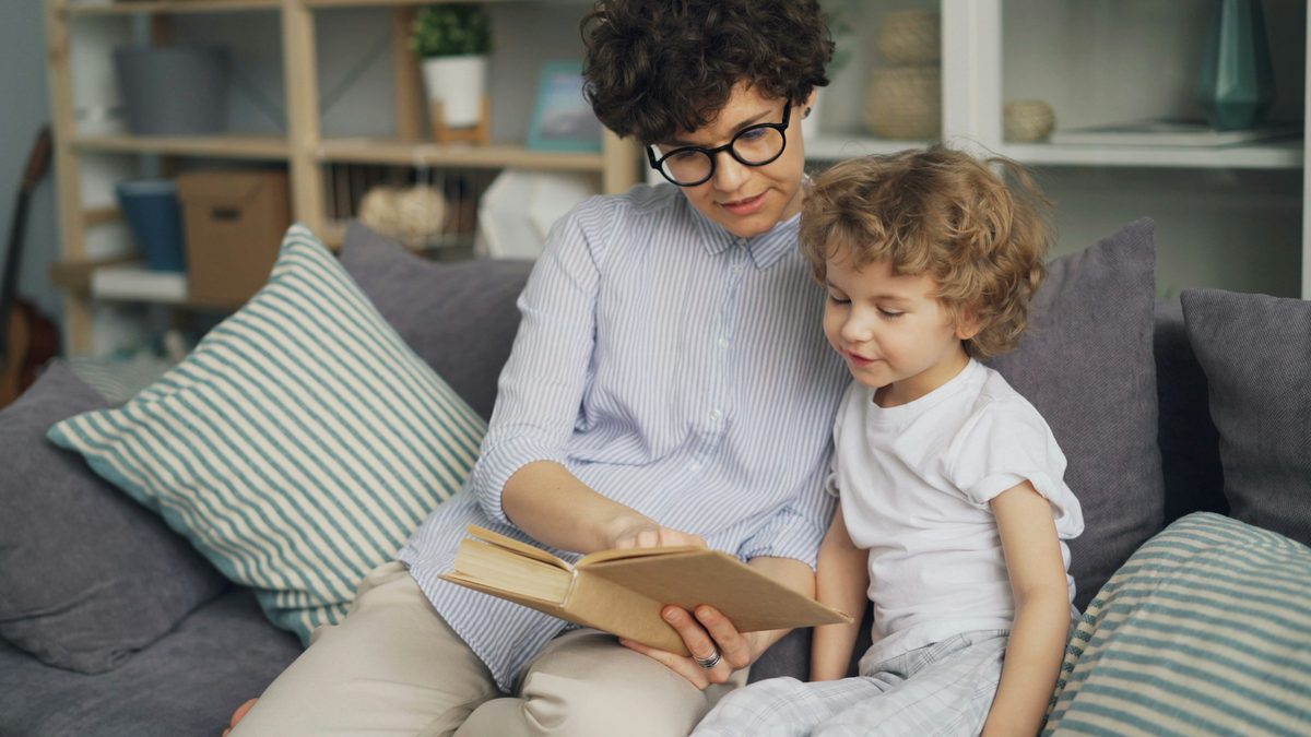 Ibu membacakan buku cerita untuk anak balita di sofa ruang keluarga