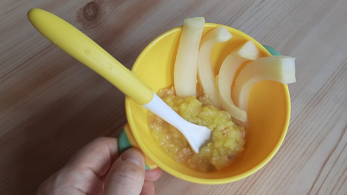 Mangkuk kuning berisi bubur MPASI buatan rumah dengan sendok bayi, dipegang orang tua di atas meja kayu